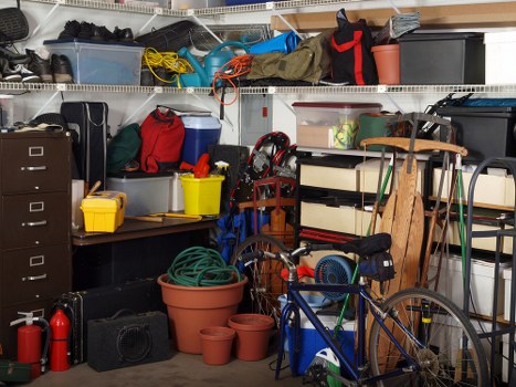 The image shows a cluttered garage or storage space filled with various household items and tools. In the foreground, there are several plant pots of different sizes and materials, some empty and stacked. To the right, a blue bicycle is leaning against a storage shelf, with a small black bag attached to its frame. Behind the bicycle, a wooden garden chair and a green garden hose are partially visible, among other miscellaneous items. The background features wire shelving units stocked with an assortment of boxes, bags, and containers, including a black toolbox, yellow plastic bins, and transparent storage boxes. There are also extension cords, a red and black backpack, and other equipment placed on the shelves. The space appears to be used for storage of outdoor and household equipment, with a concrete or similar plain flooring visible at the base. The overall lighting suggests an indoor environment, typical of a residential garage or utility area used for storing miscellaneous items and tools that might be considered for rubbish removal by Commercial Waste Removal Archway.