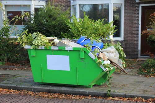 A large, bright green plastic waste skip situated on a paved driveway outside a residential brick house in Archway, North London, with white-framed windows and a brown door in the background. The skip is filled to the brim with a mixture of garden waste, including branches with green leaves, and household rubbish such as cardboard boxes, plastic bags, and miscellaneous debris. Some of the waste is spilling over the sides, resting on the pavement, with a few scattered leaves and twigs nearby. The surrounding environment features a well-maintained garden with dense shrubbery and small trees behind the skip, and a curb running along the edge of the driveway. This scene visually represents a typical rubbish removal process in an urban residential setting, aligning with the services offered by Commercial Waste Removal Archway in the local postcode area.
